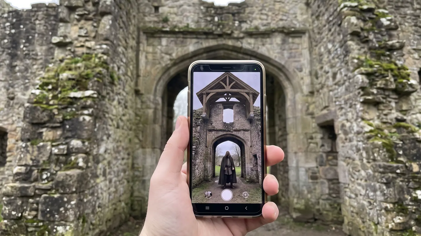 A phone held up to a ruined gatehouse shows the arch rebuilt with its timber roof and a hooded figure standing beneath